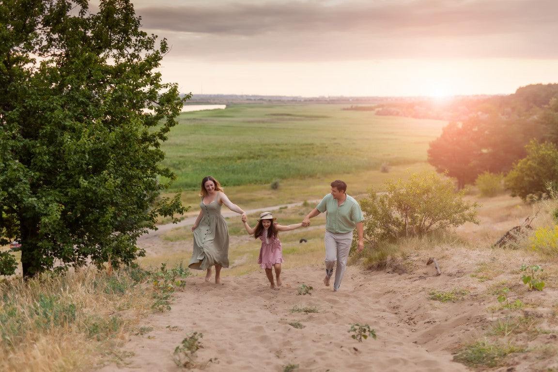 Eine glückliche Familie spaziert barfuß durch den Sand auf einem Hügel in der Natur, während die Sonne untergeht.
