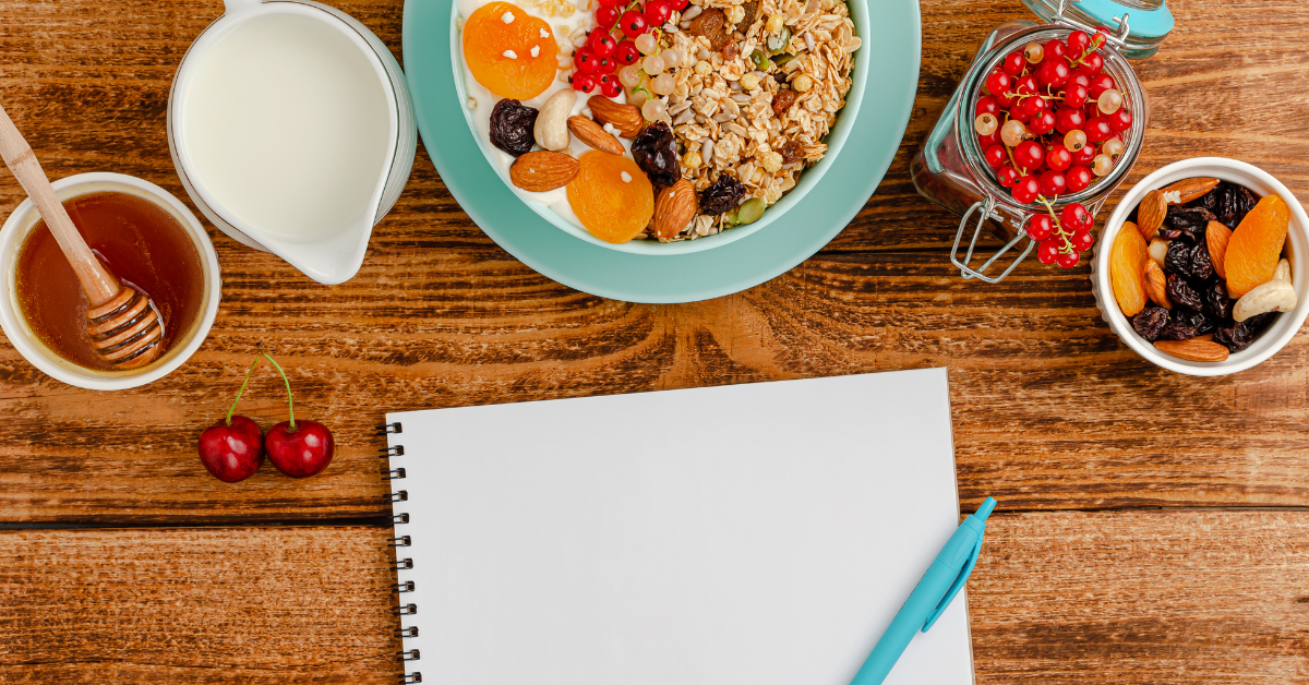 Gesundes Frühstück mit Müsli, Joghurt, Nüssen und Obst neben einem leeren Ernährungstagebuch auf einem Holztisch.