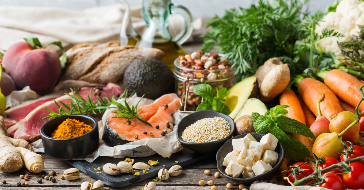 Auswahl an Lebensmitteln mit Lachs, Avocado, Pistazien, Pflanzenöl, Brot, Hülsenfrüchten sowie frischem Obst und Gemüse.