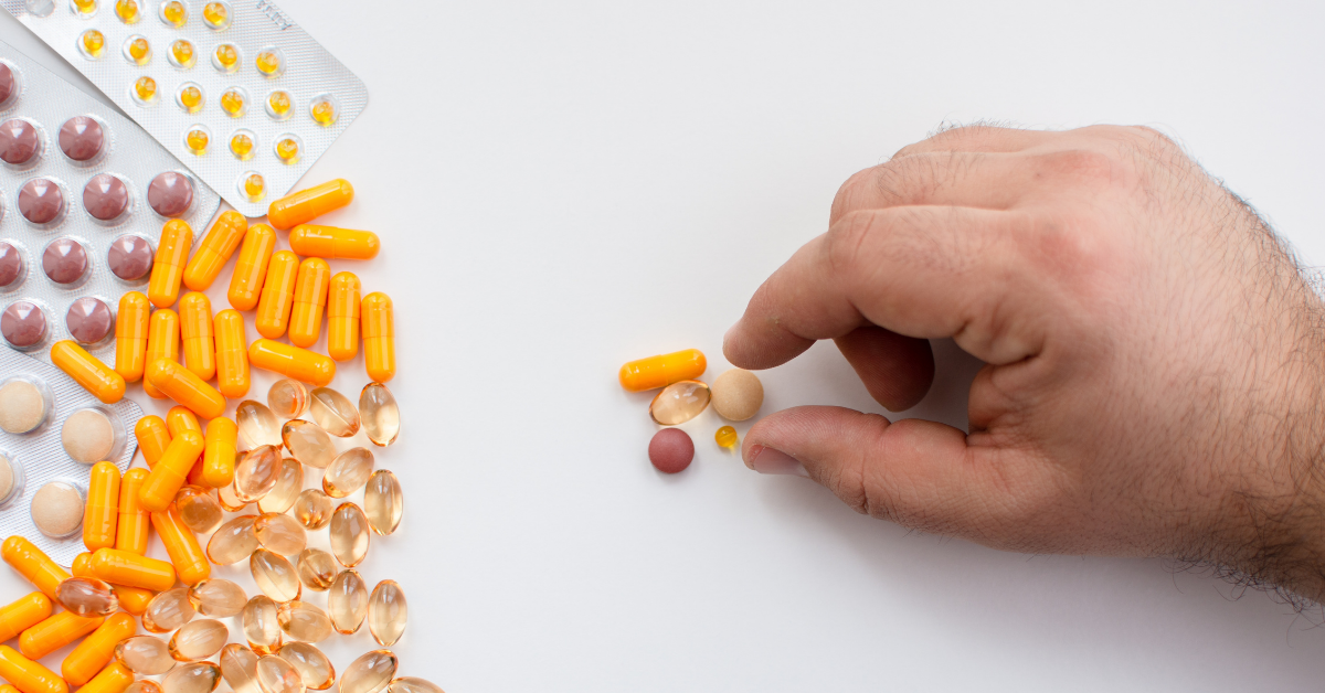 Hand greift gezielt einzelne Kapseln und Tabletten aus einer Vielzahl von Nahrungsergänzungsmitteln – Symbol für bewusste Dosierung von Supplements