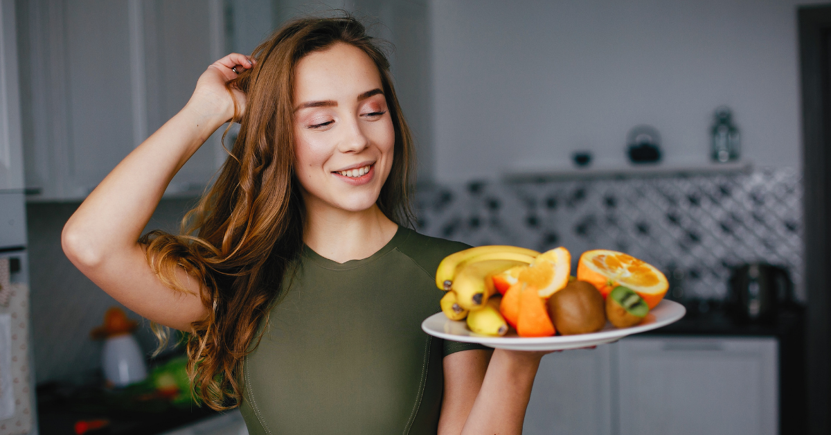 Junge Frau hält einen Teller mit frischem Obst wie Bananen, Orangen und Kiwi in der Hand und lächelt.