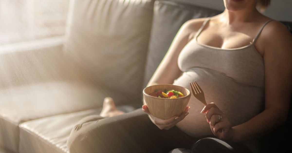 Schwangere Frau sitzt auf dem Sofa und genießt eine ausgewogene Mahlzeit – Ernährung während der Schwangerschaft.
