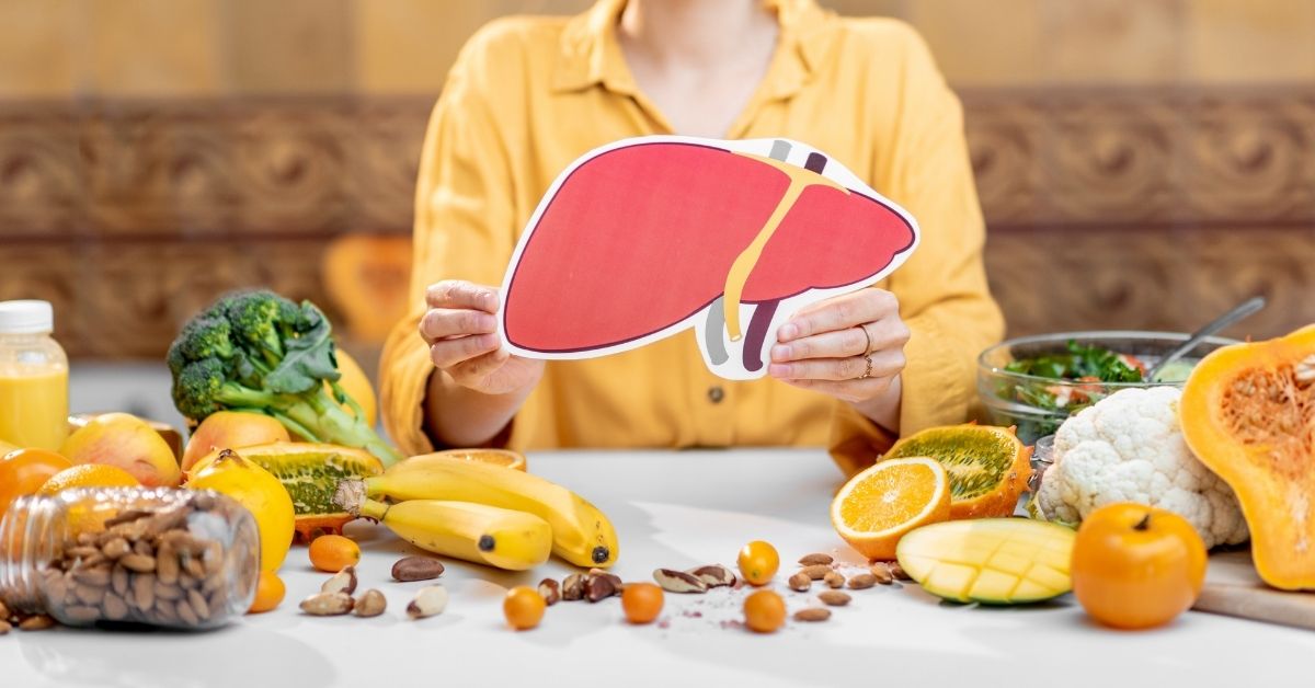 Frau hält ein Lebermodell zwischen Obst, Gemüse, Nüssen und weiteren Lebensmitteln für eine leberfreundliche Ernährung.