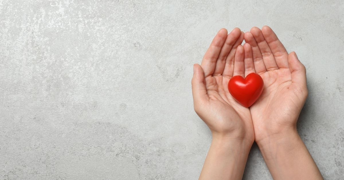 Hände halten ein kleines rotes Herz aus Holz – Symbol für Herzgesundheit, Vorsorge und kardiovaskuläres Wohlbefinden.