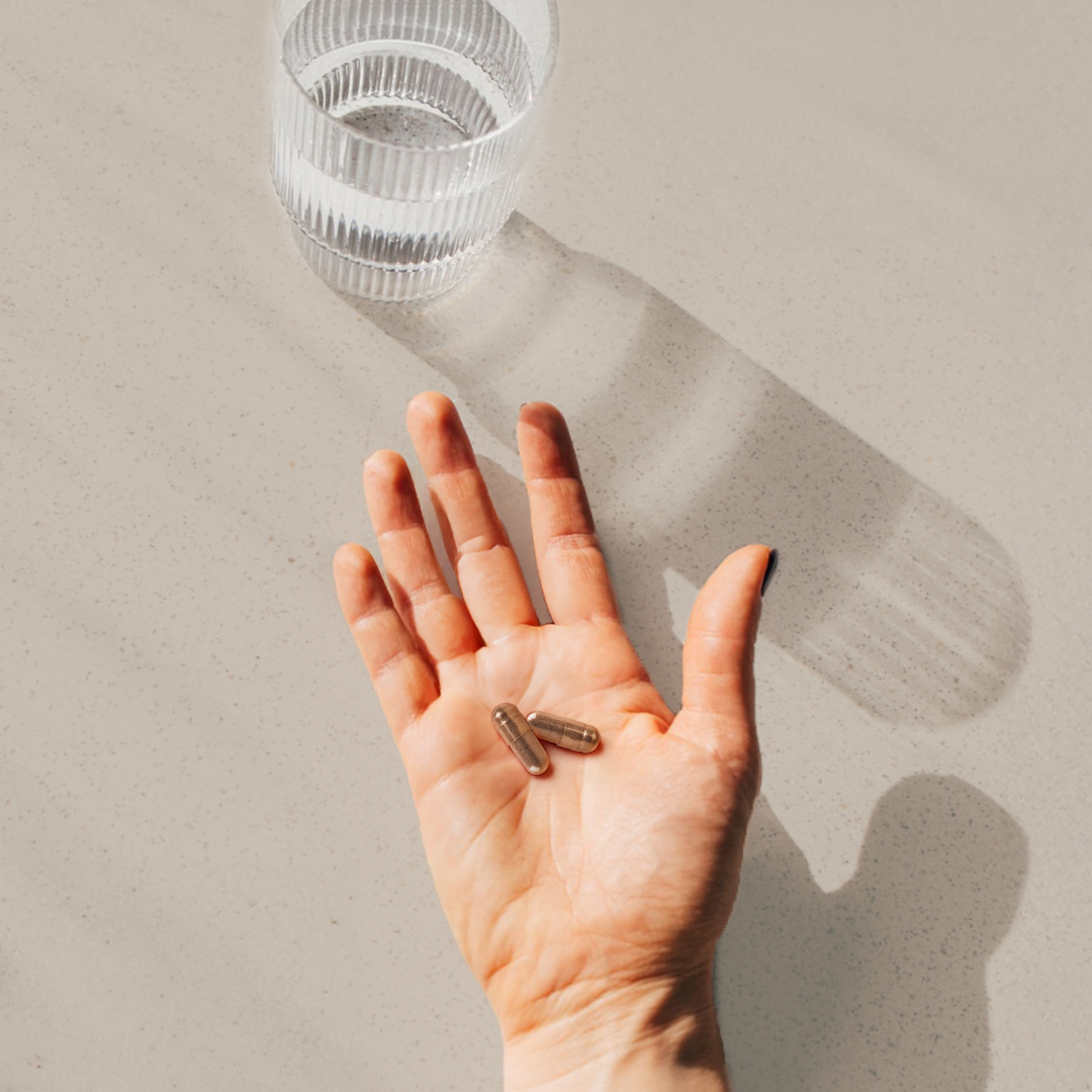 Hand, die 2 braune Kapseln hält, daneben ein Wasserglas