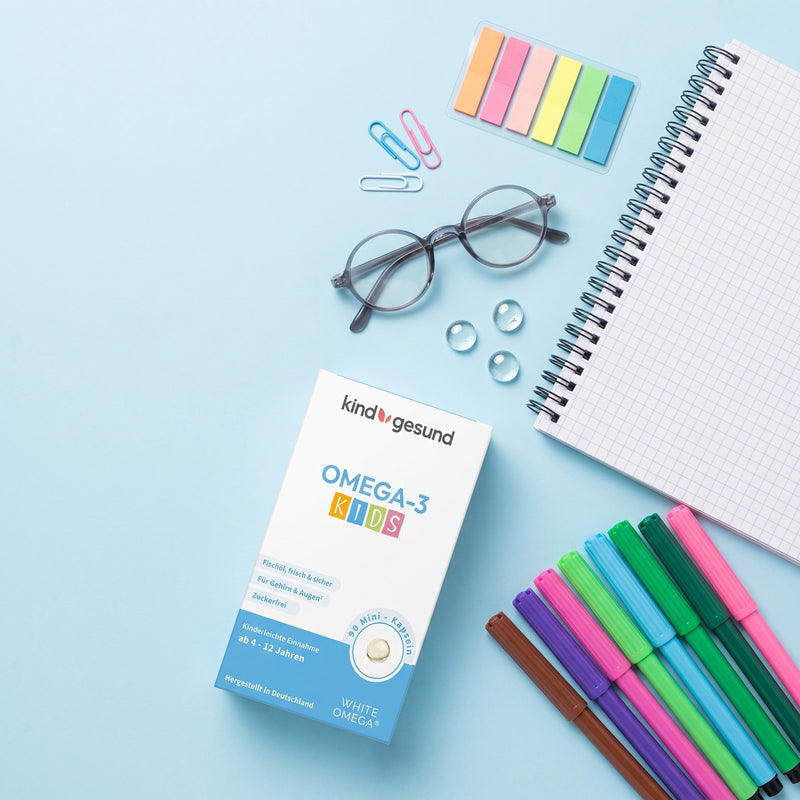 kindgesund Omega-3 KIDS Mini-Kapseln neben Lesebrille, kariertem Notizblock, bunten Filzstiften und Haftmarkern auf hellblauem Untergrund