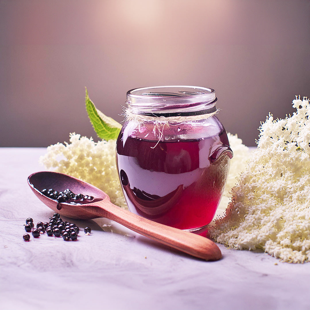 Tiefviolette Beeren des Holunders auf einem Löffel neben einem gefüllten Glas und Holunderblüten